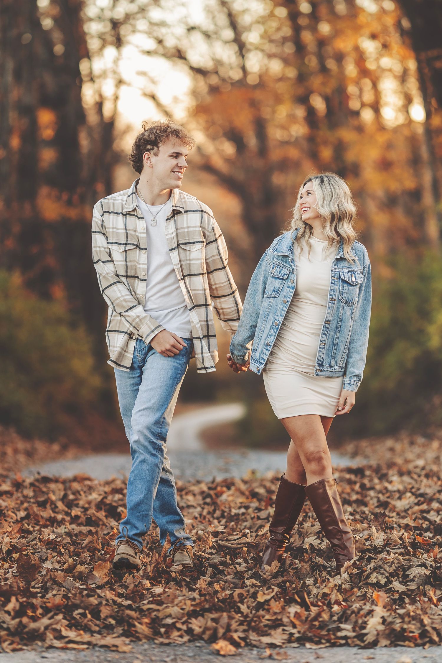 Couple holding hands on a leaf-covered path surrounded by autumn foliage, wearing casual fall outfits.