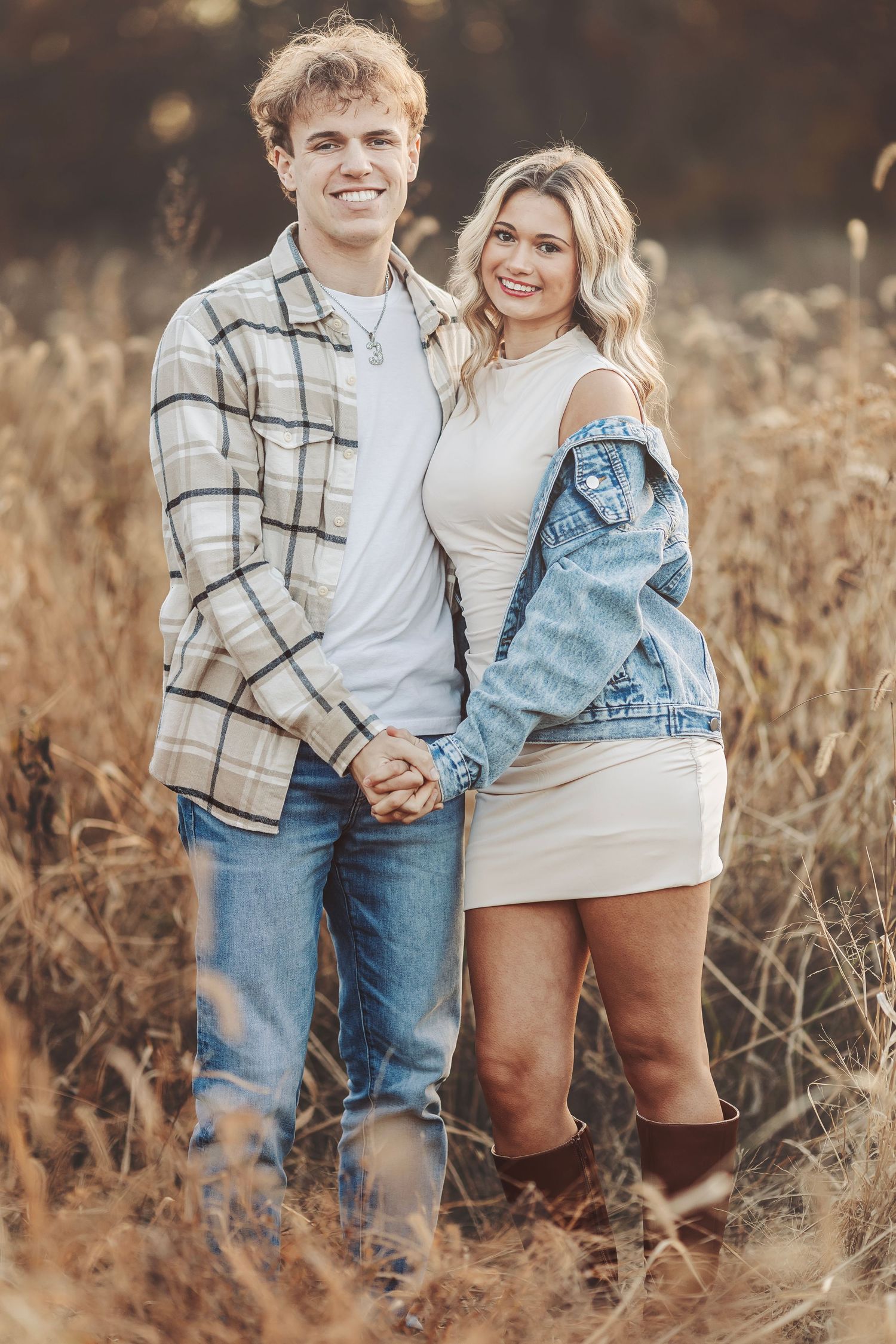 A smiling young couple holds hands in a golden field, the man in plaid and the woman in a white dress with denim jacket.