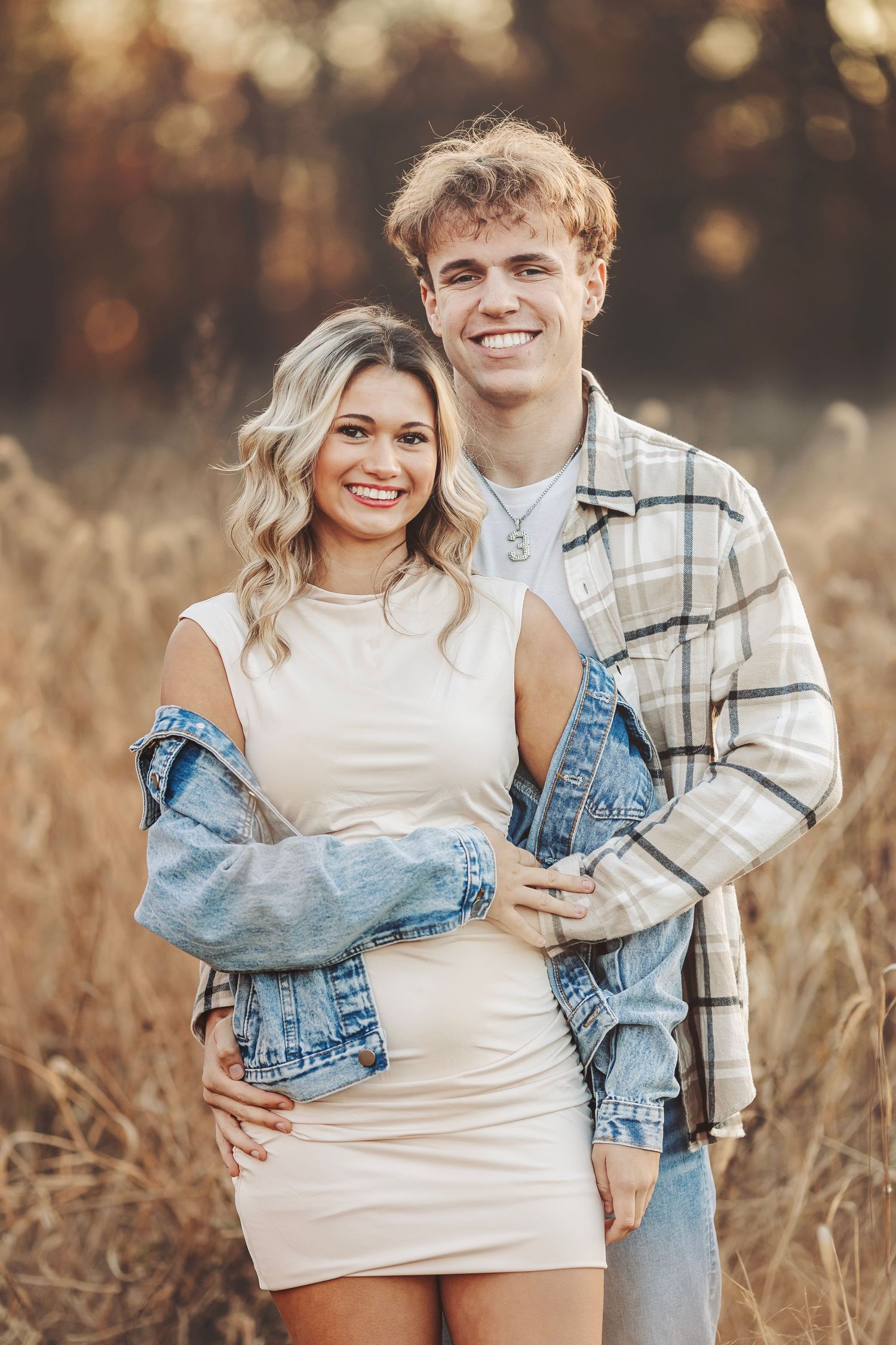 A smiling young couple poses together in a golden field, she in a cream dress with denim jacket, he in a plaid shirt.