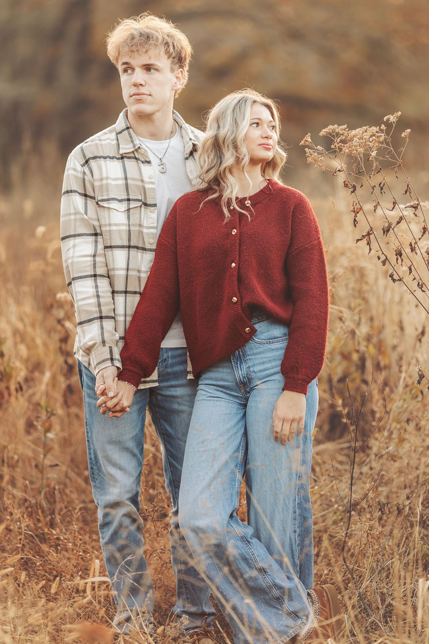 Young couple holding hands in autumn field, wearing jeans and cozy sweaters, surrounded by golden dry grass.