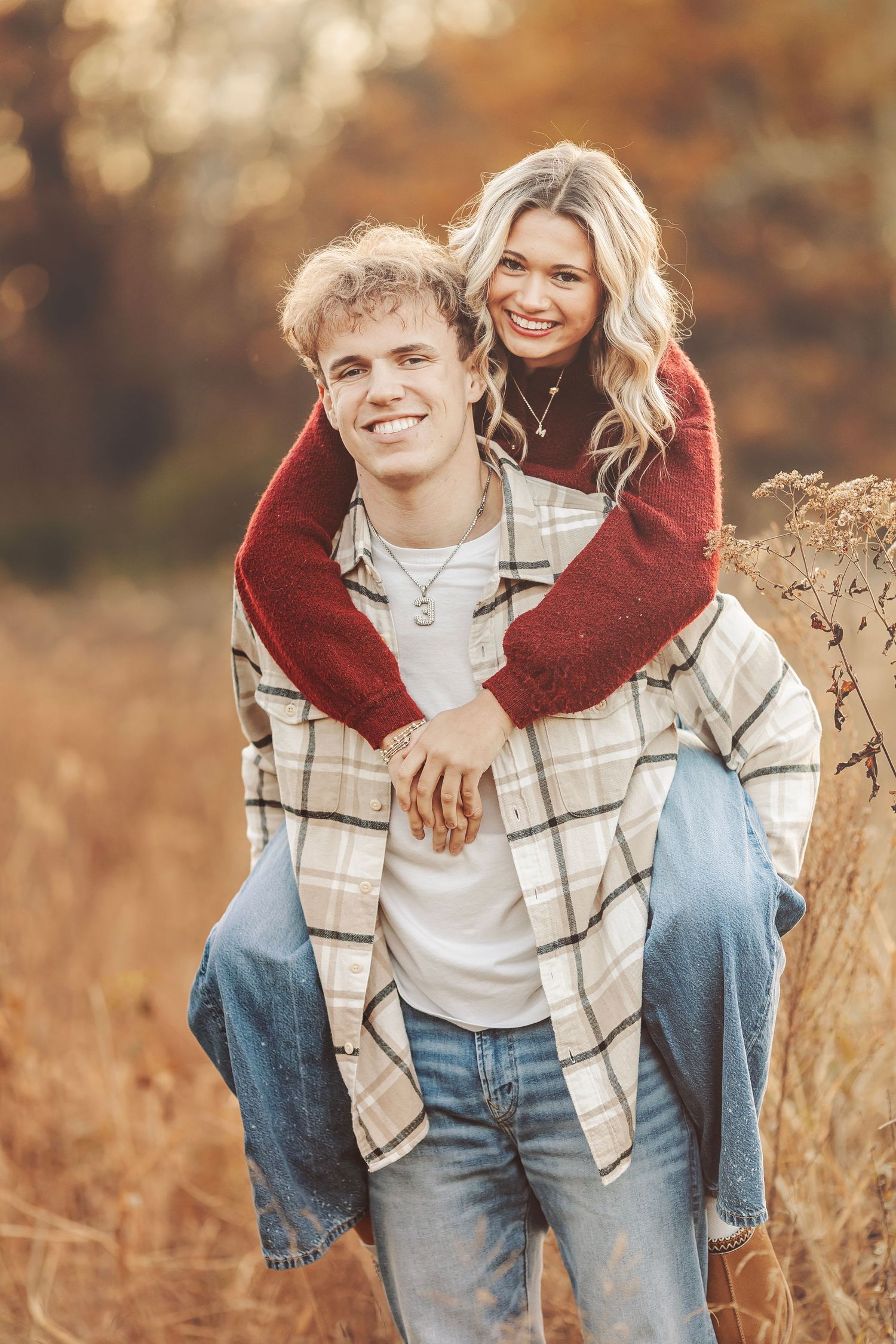 A young couple poses outdoors in fall, girl in red sweater piggyback riding a guy in plaid shirt, both smiling.