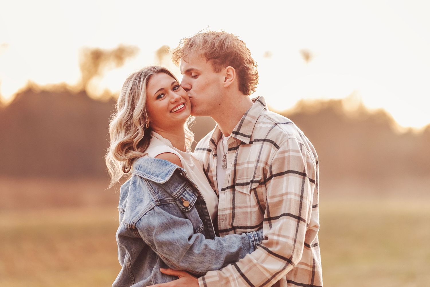 A couple shares a romantic sunset kiss outdoors, the man in plaid and woman in denim jacket embracing in golden light.