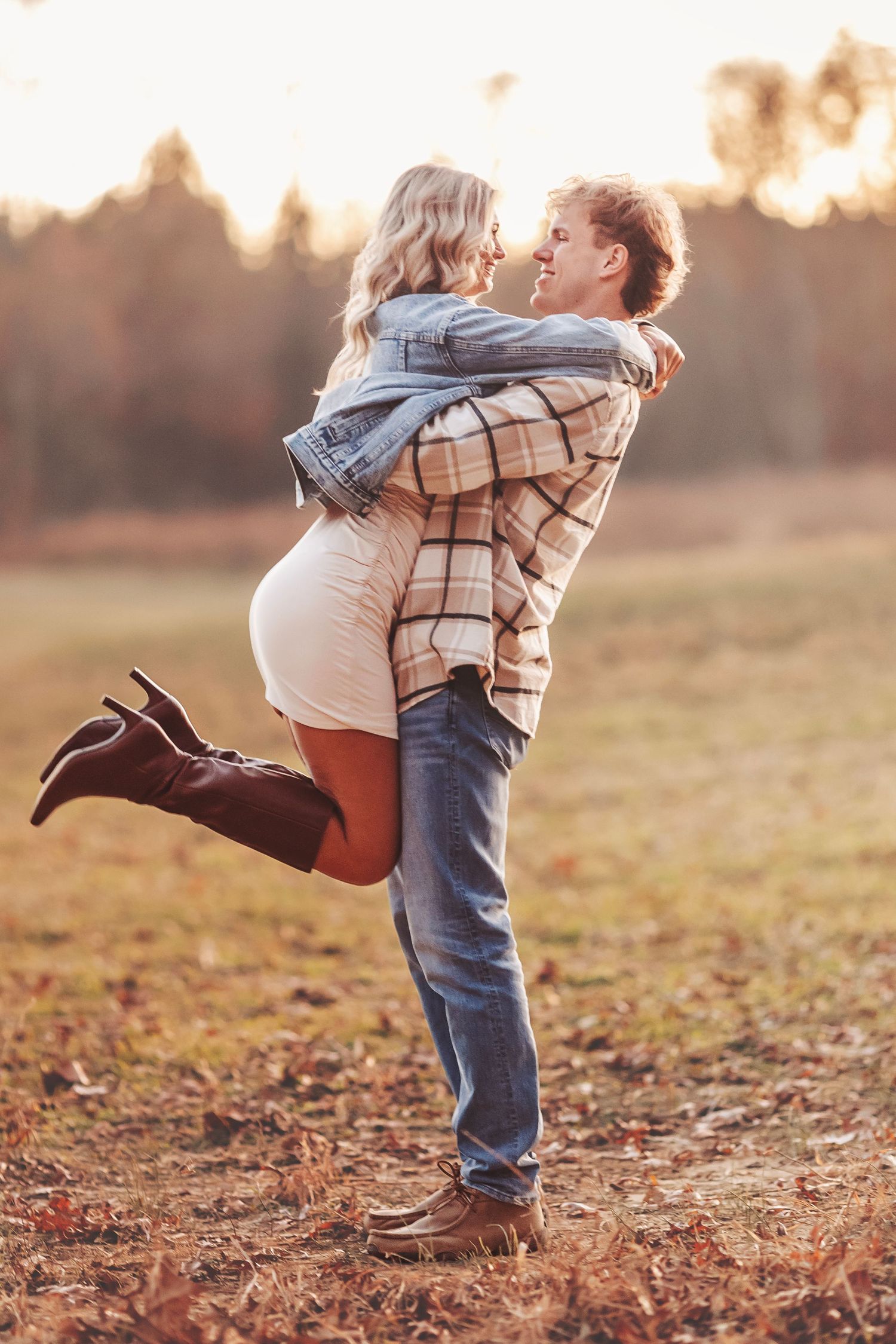 Couple embracing in autumn field, man lifting woman in plaid jacket and boots during golden hour.