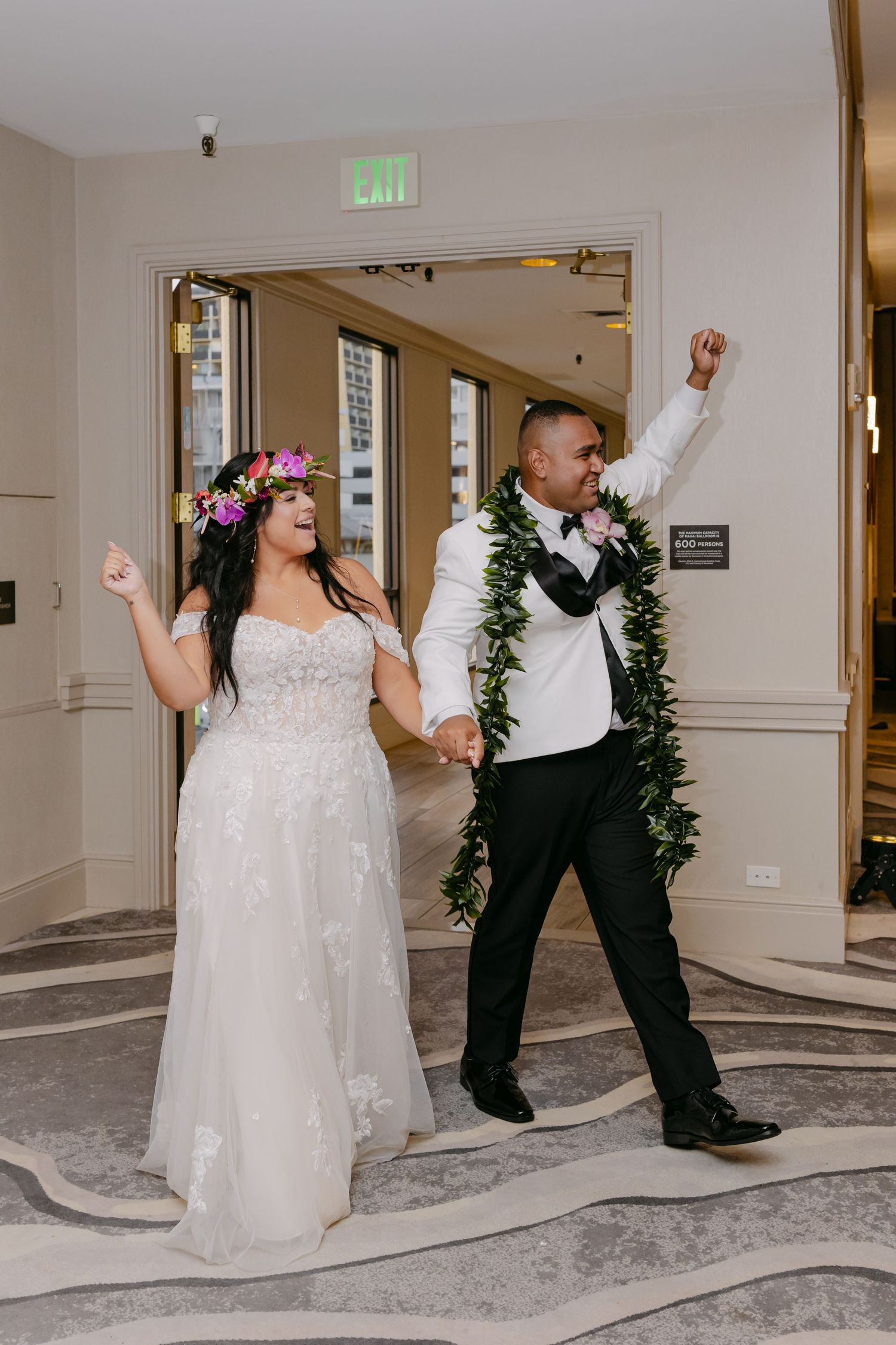 Joyful newlyweds make grand entrance, bride in floral crown and gown, groom in tuxedo with green lei, arms raised.