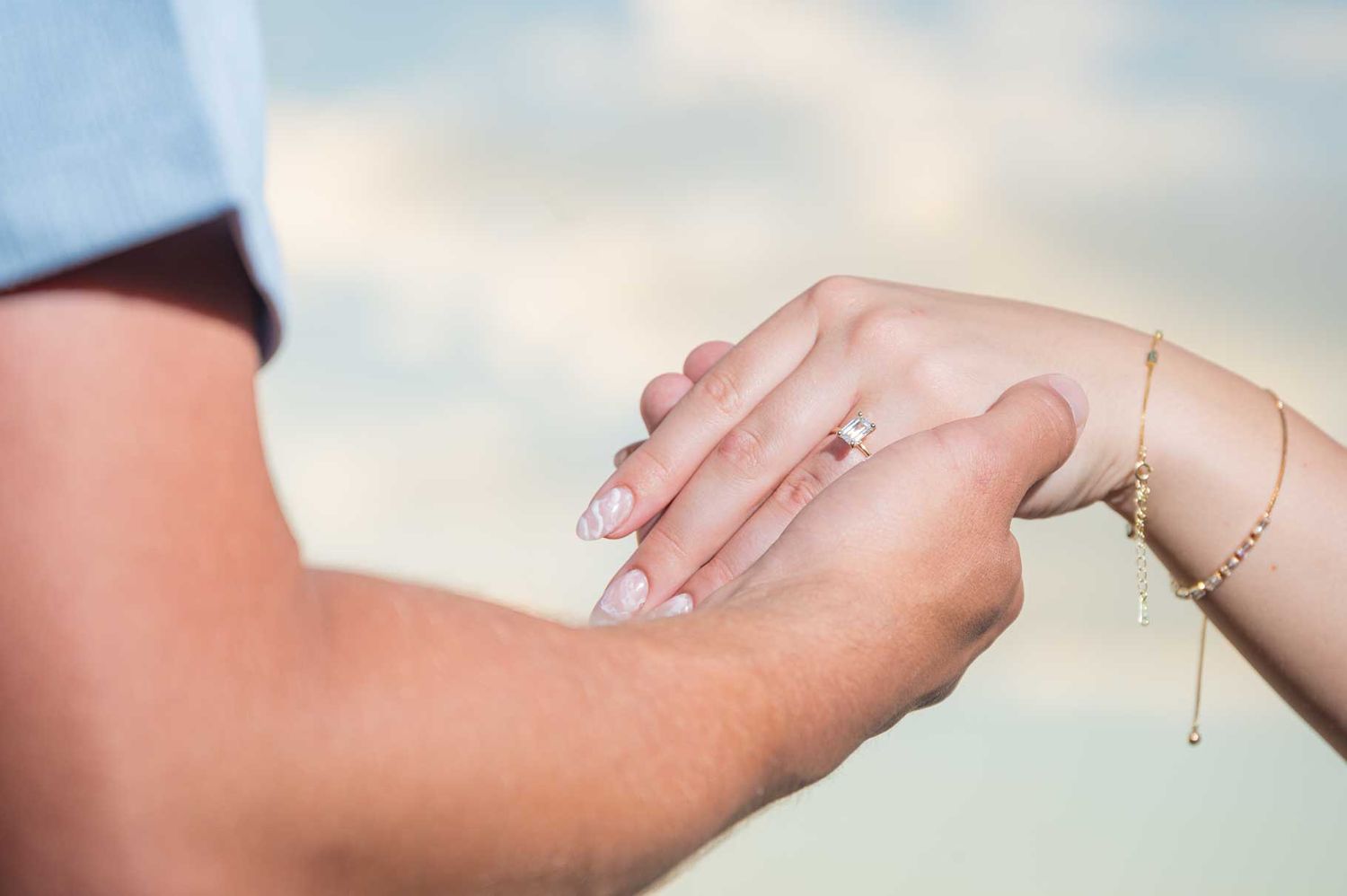 Close-up of a couple holding hands with an engagement ring on the woman's finger against a soft sky background.