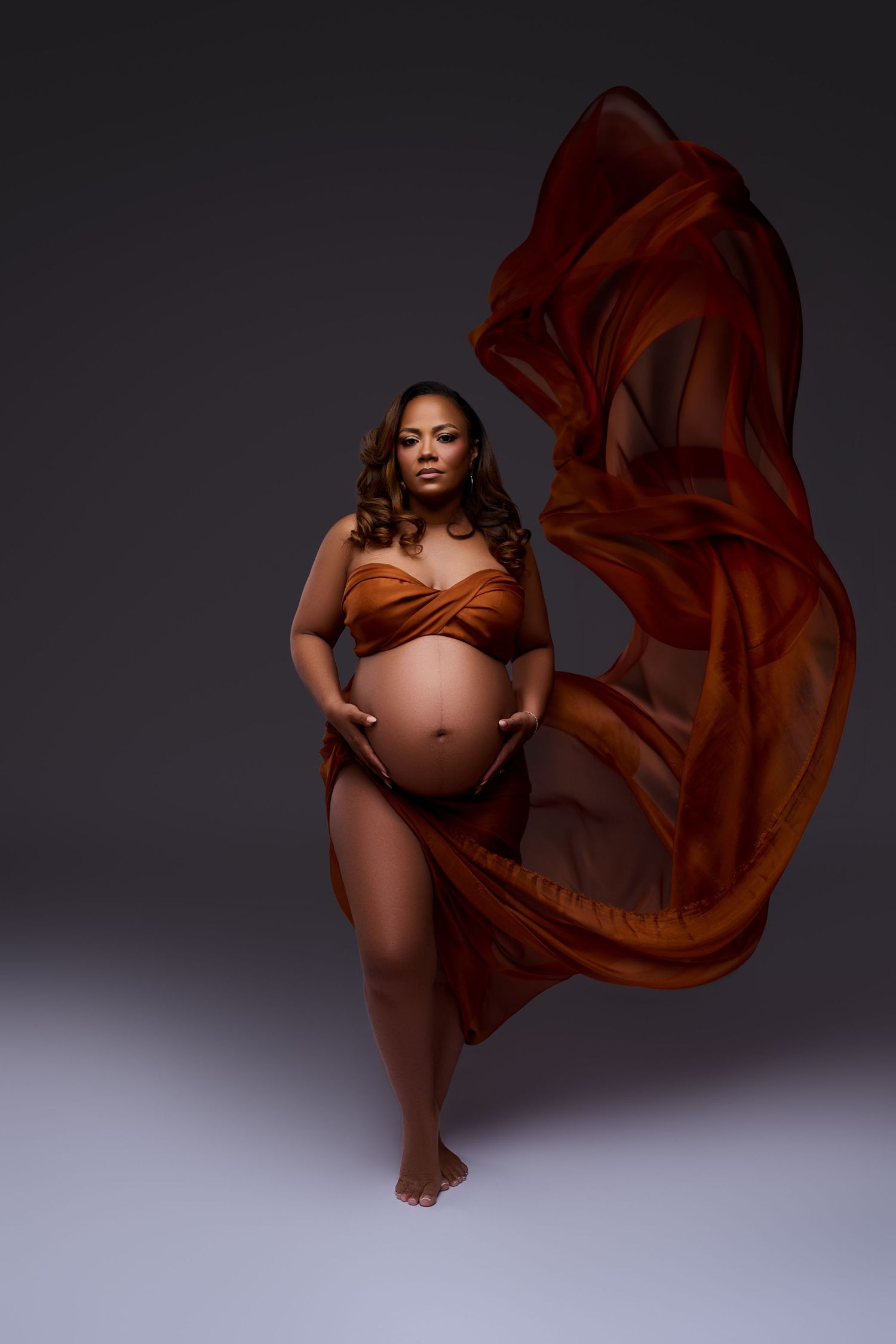 Pregnant woman in flowing rust-orange fabric draped elegantly, posing against gray studio backdrop in artistic maternity portrait.