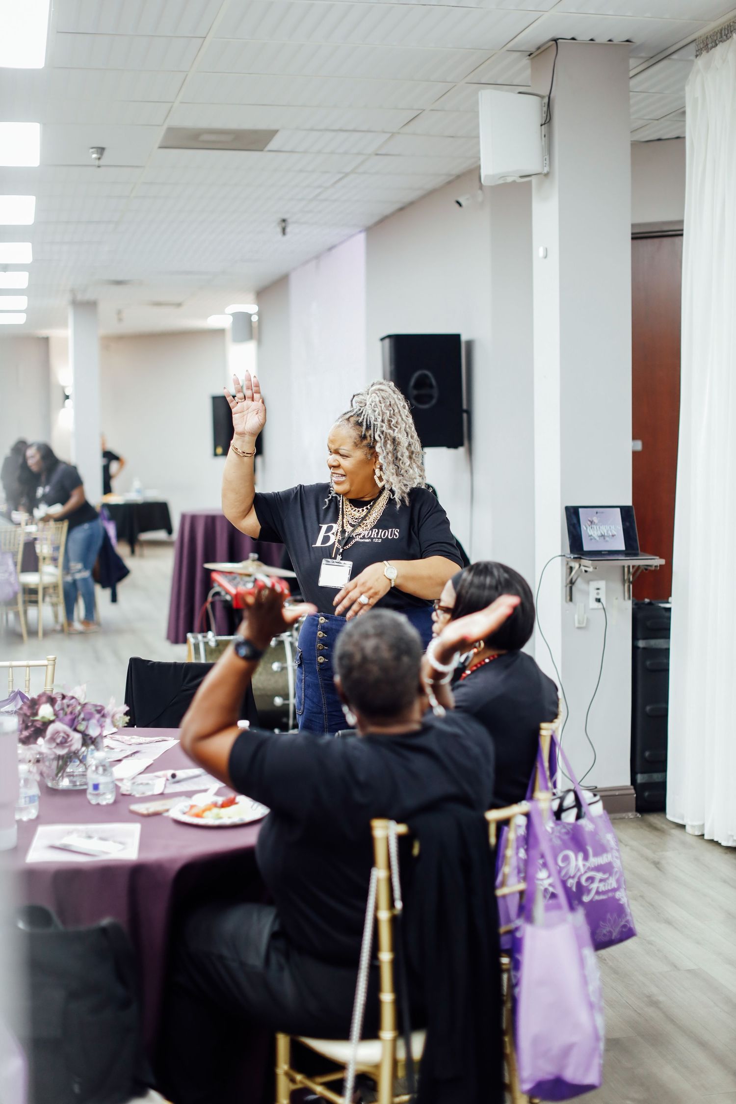 Woman with gray locs raises hand at purple-themed banquet event while seated guests listen attentively in decorated hall.