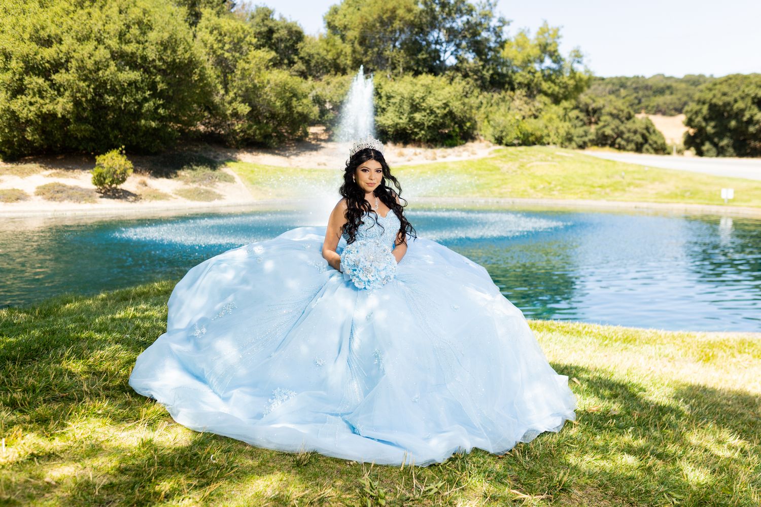 Young woman in a voluminous light blue quinceañera ball gown poses by a fountain and pond in a lush green park.