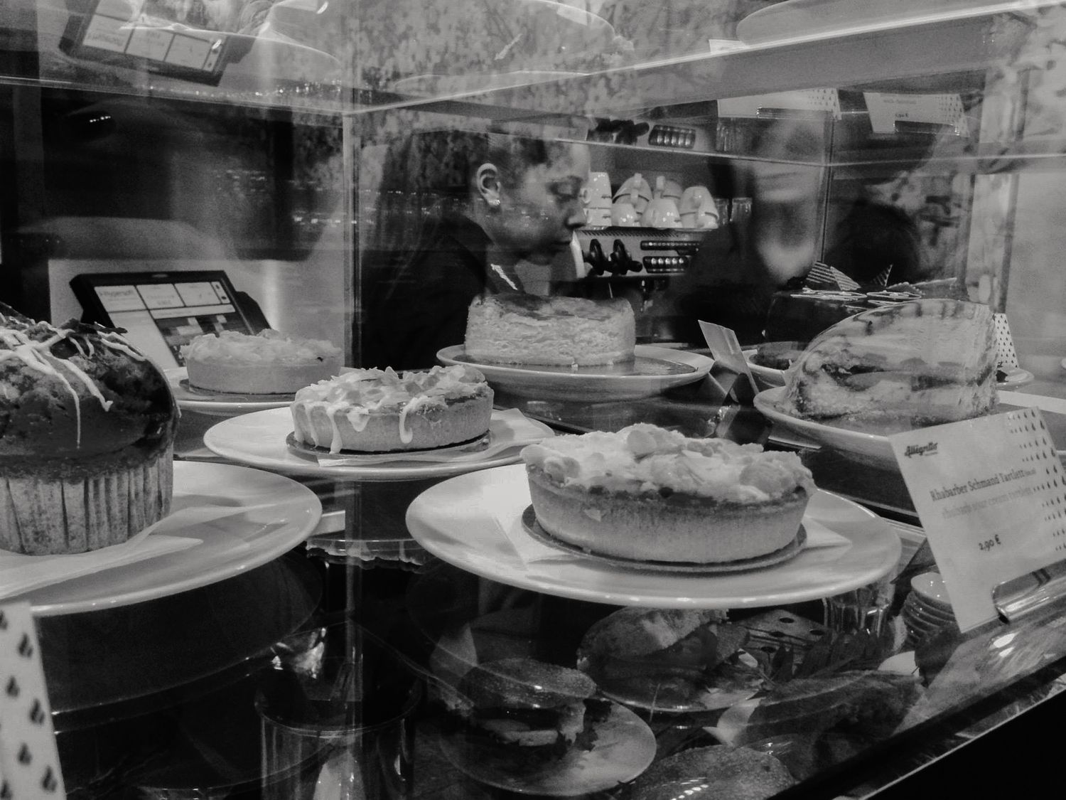 Black and white bakery display case with assorted cakes and couple reflected in glass window.