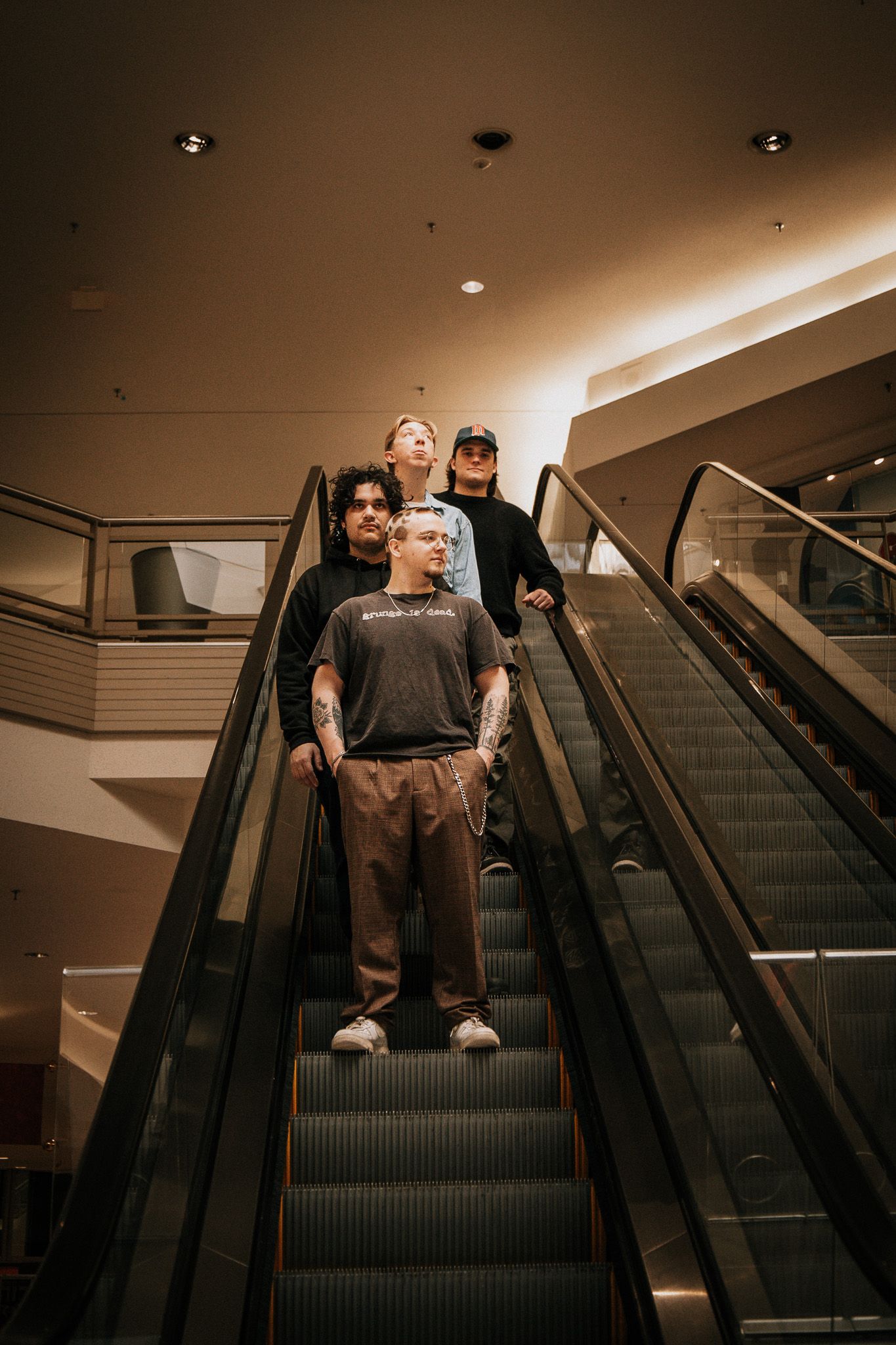Three young people posing on an escalator in a mall, wearing casual streetwear and tattoos.