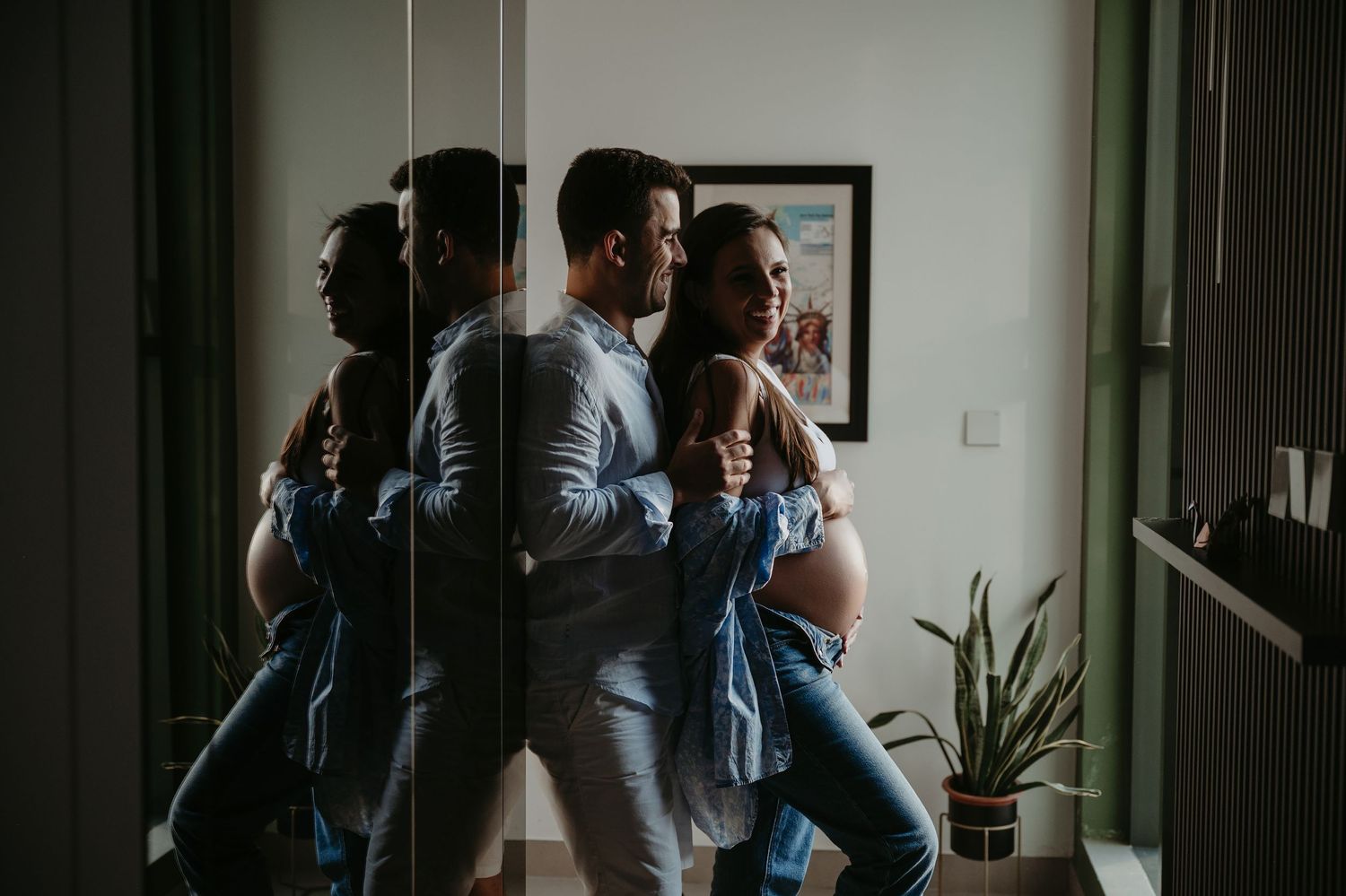 Couple in denim leaning against mirror wall, woman with baby bump, soft indoor light, maternity photo session.