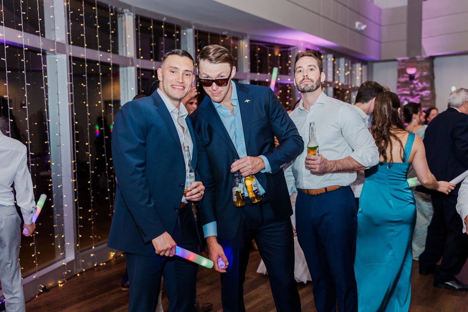 Three men in suits posing at a party with glow sticks and drinks, with fairy lights in the background.