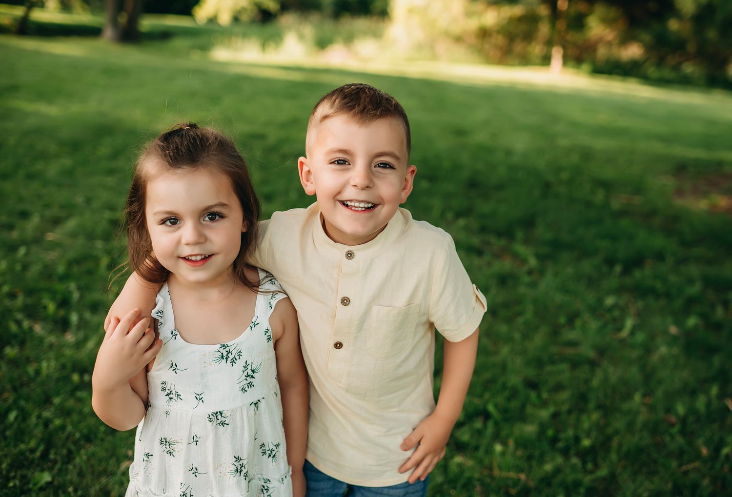 Two happy young siblings smiling outdoors on a sunny day in a lush green park, girl in floral dress and boy in beige shirt.