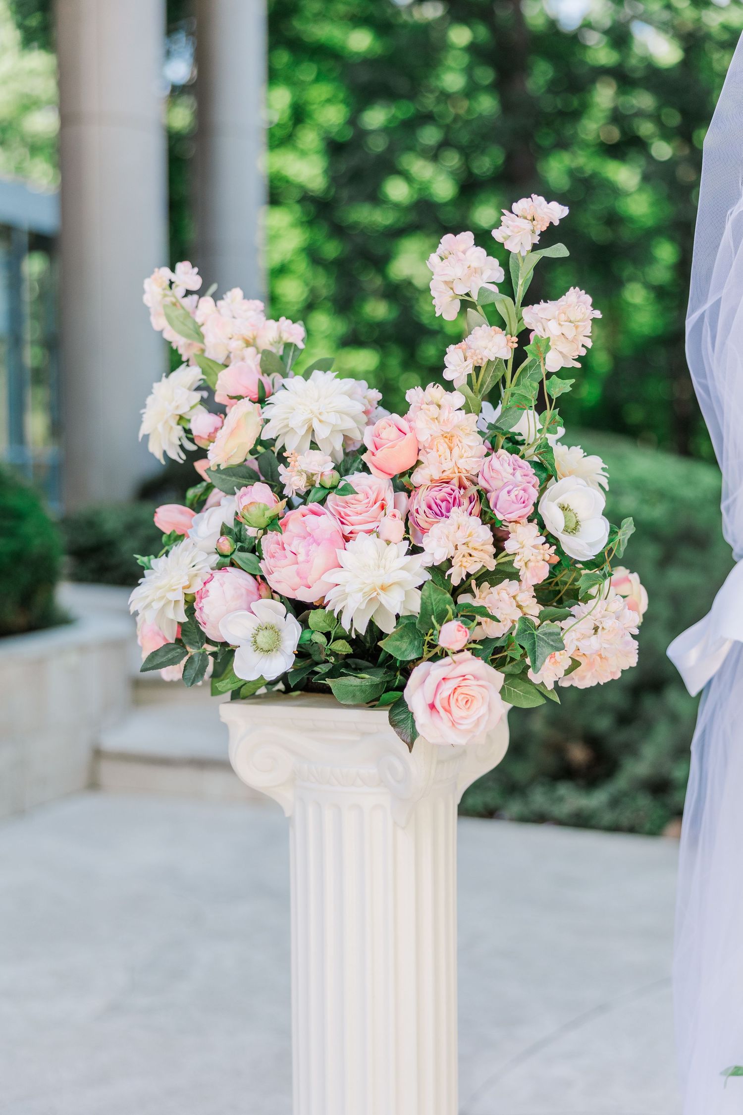 Elegant white pedestal column adorned with a lush arrangement of pink and white roses, peonies, and dahlias at an outdoor wedding.