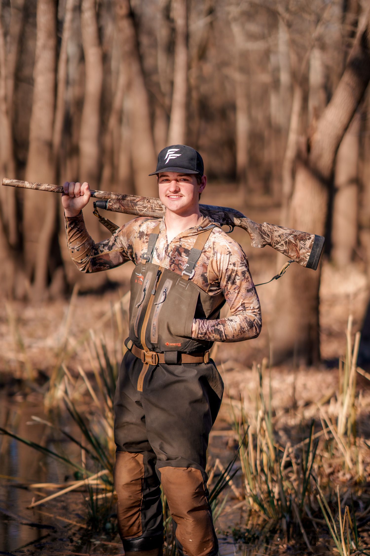 Smiling hunter in camo gear carrying a rifle over his shoulder while standing in a wooded wetland area.