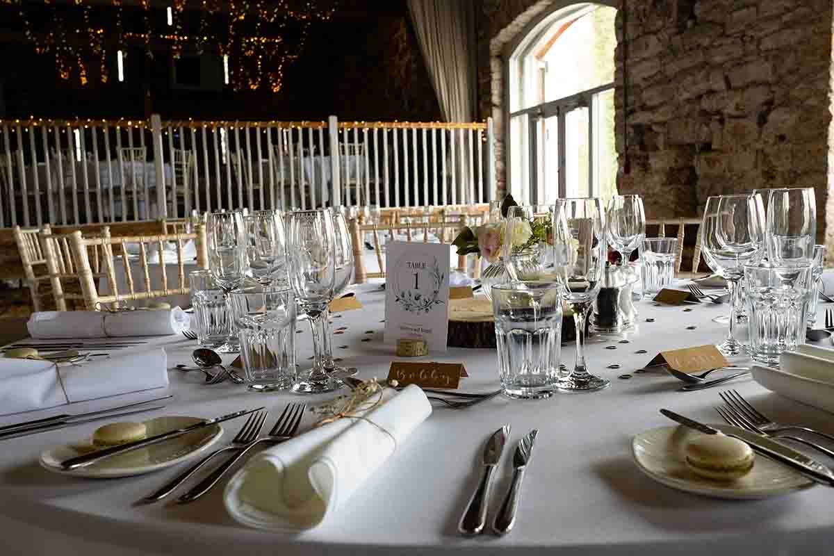 Elegant wedding reception table set with crystal glasses, silverware, and white linens in a rustic stone venue.