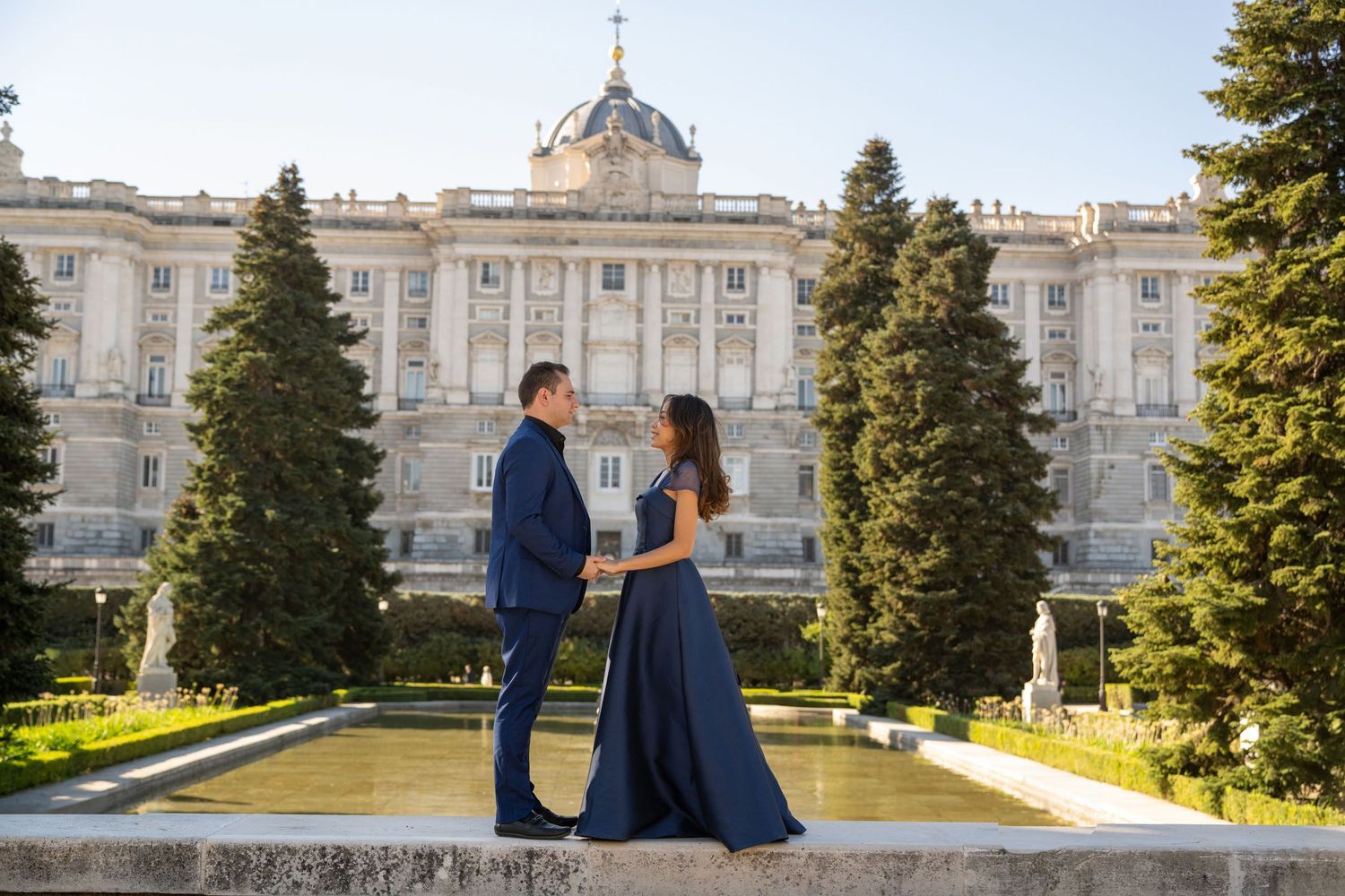 Couple in elegant navy attire posing in front of the Royal Palace of Madrid gardens with fountains.