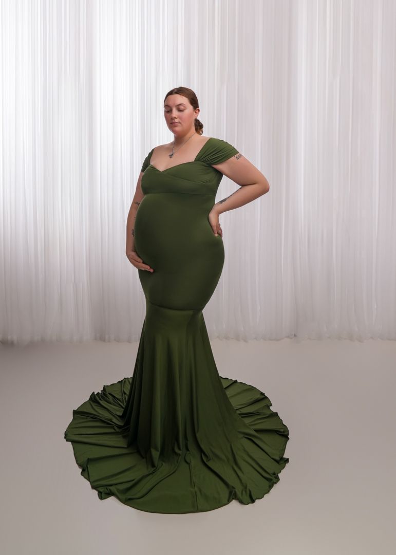 Pregnant woman in elegant olive green mermaid gown with off-shoulder design and long train, posing gracefully.