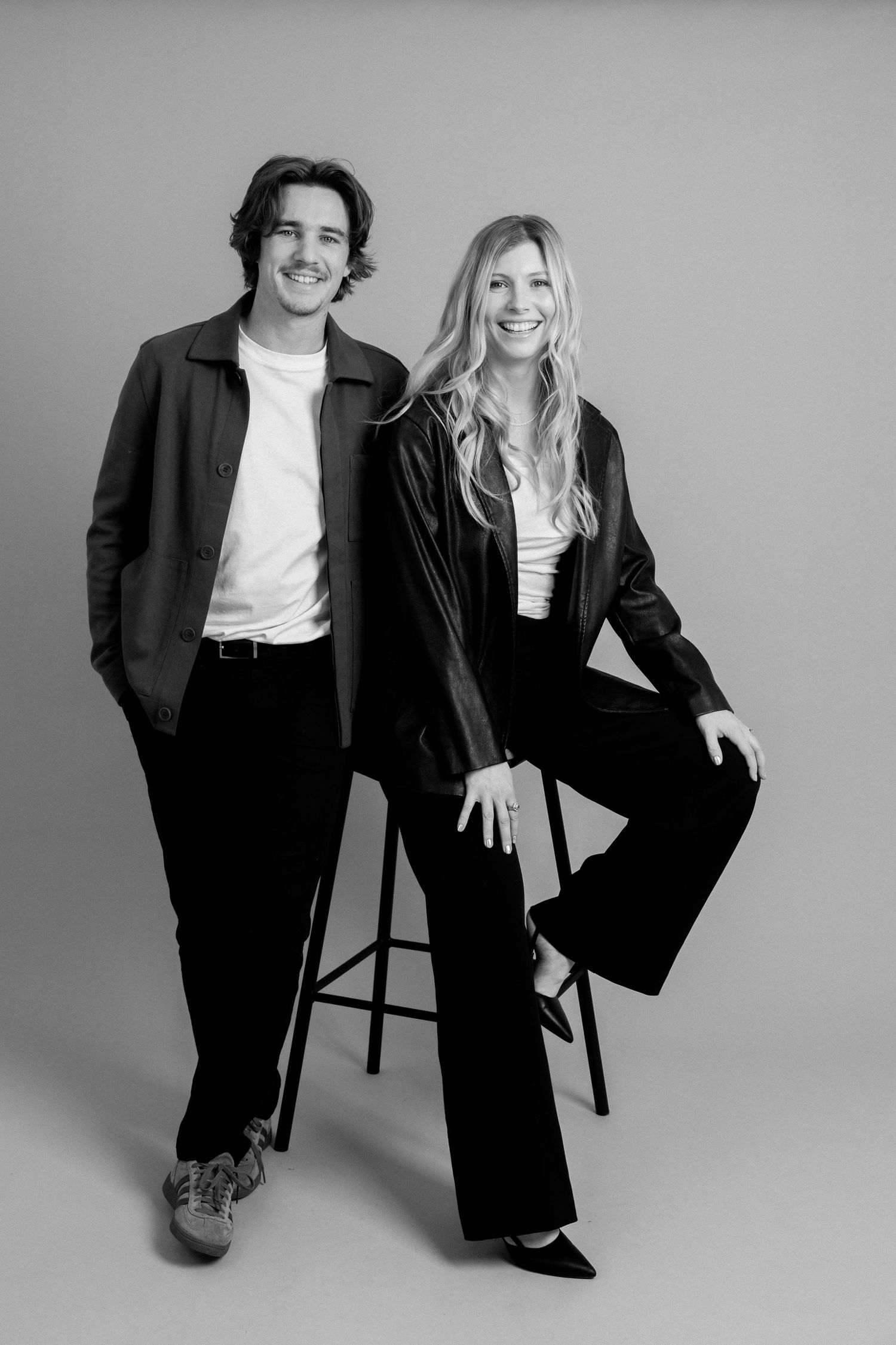 Black and white studio portrait of a smiling man and woman in dark blazers posing together against a neutral background.
