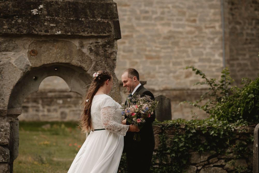Bride and groom sharing a romantic moment outside a historic stone church, bride holding a floral bouquet.