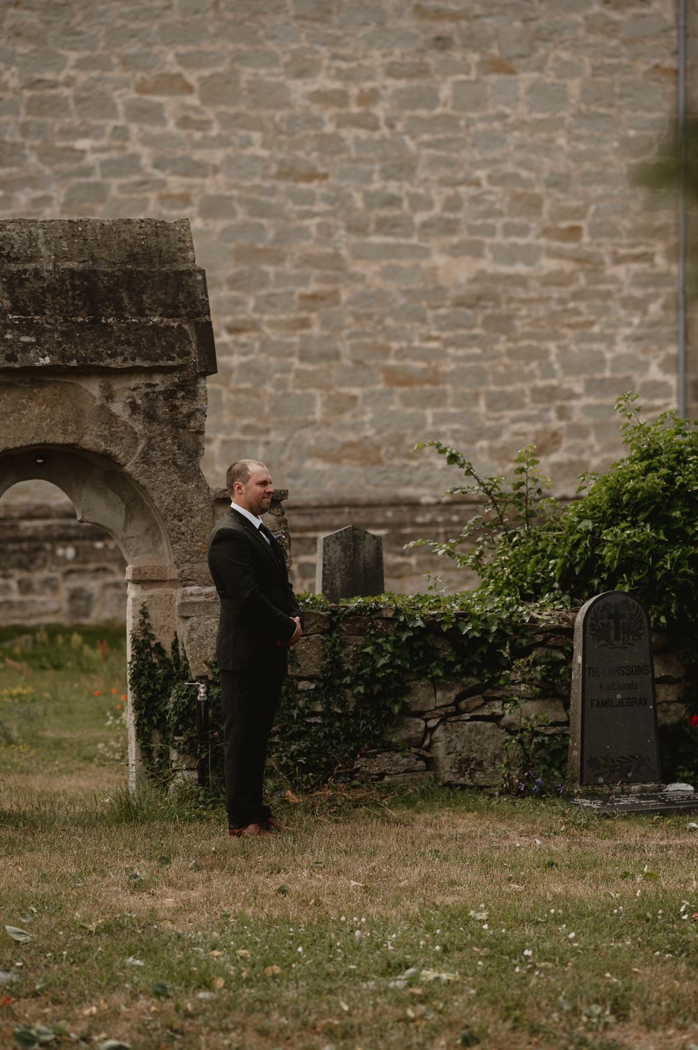 A groom in a black suit stands contemplatively among ancient stone grave markers beside a historic church wall.