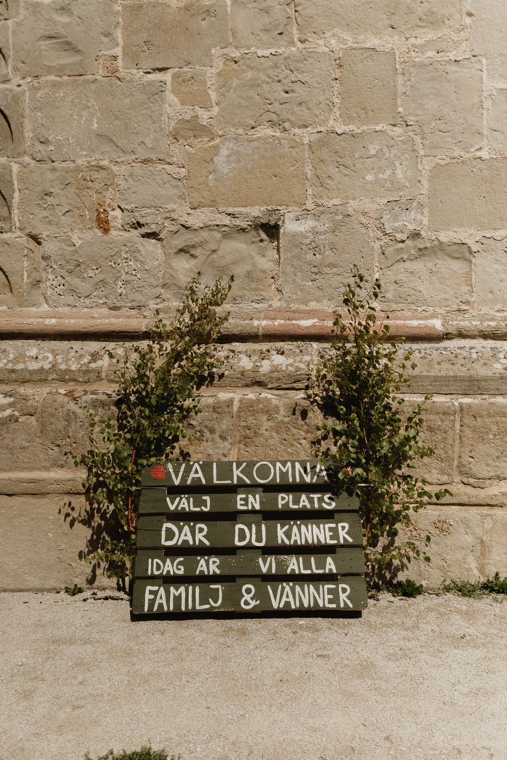 Swedish wedding welcome sign reading Valkomna with greenery against a rustic stone wall.