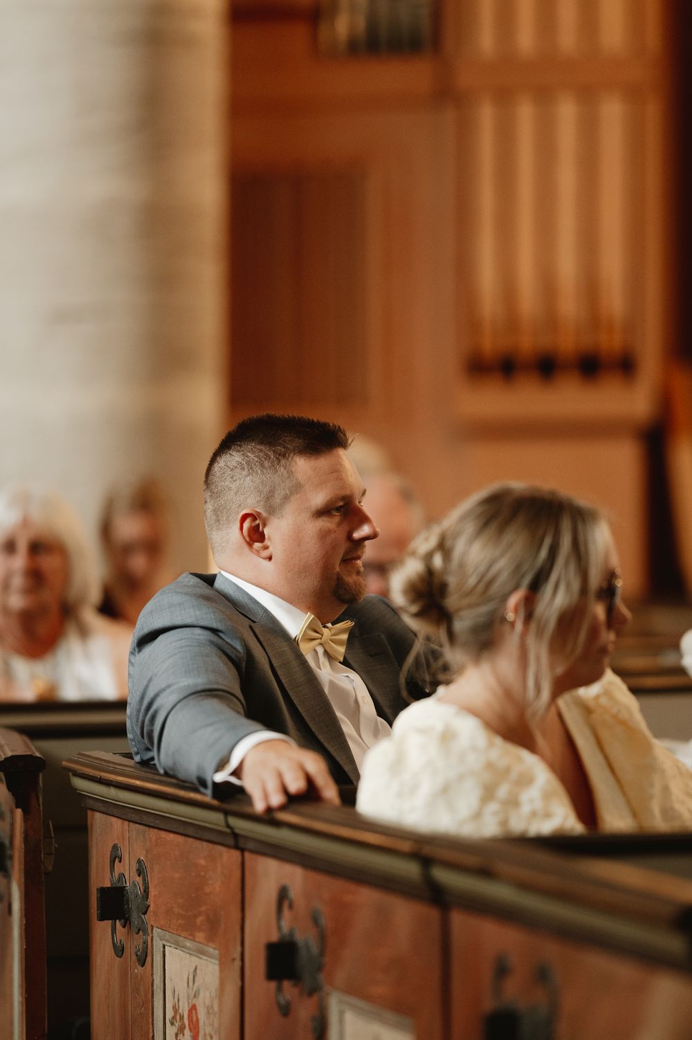 A groom in a gray suit and gold bow tie sits in a church pew during a wedding ceremony.