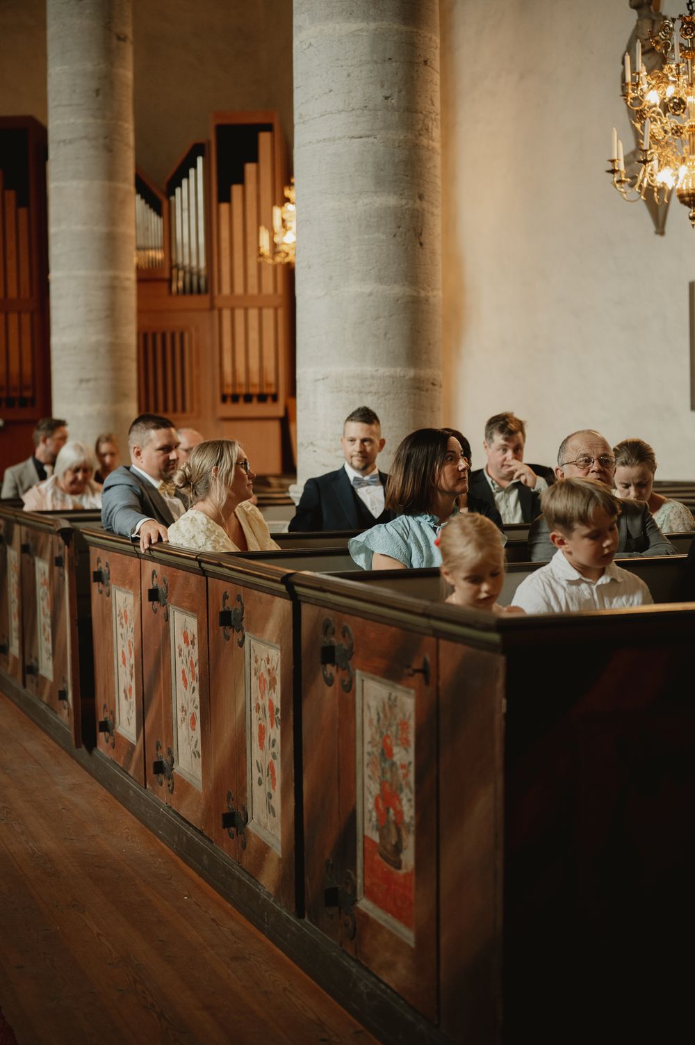 Guests seated in ornate wooden church pews during a wedding ceremony, with a pipe organ visible in the background.