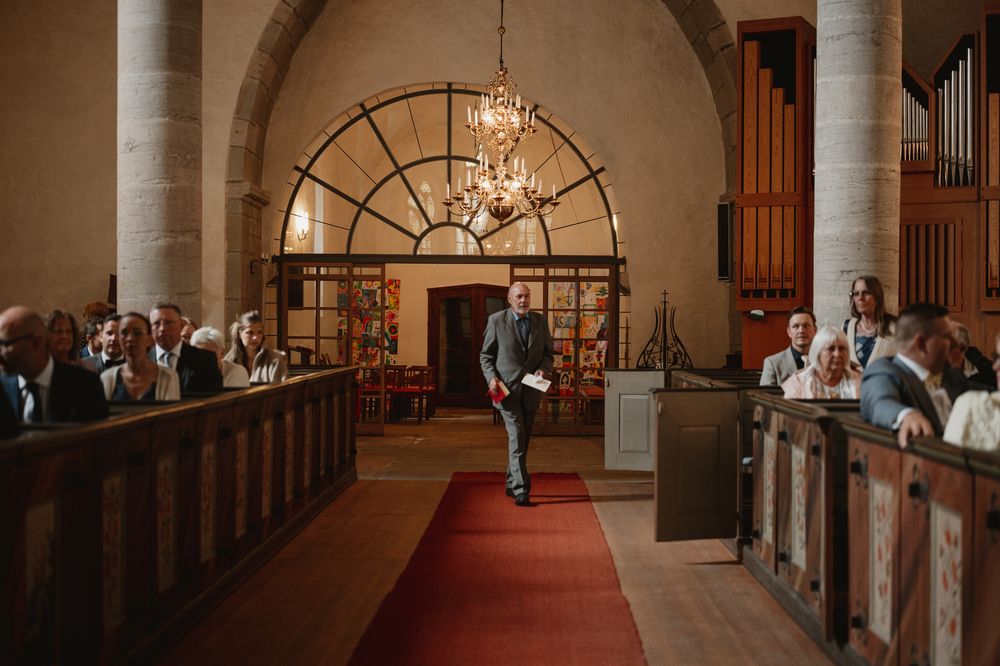 Groom walks down red carpet aisle in historic stone church surrounded by wedding guests during ceremony.