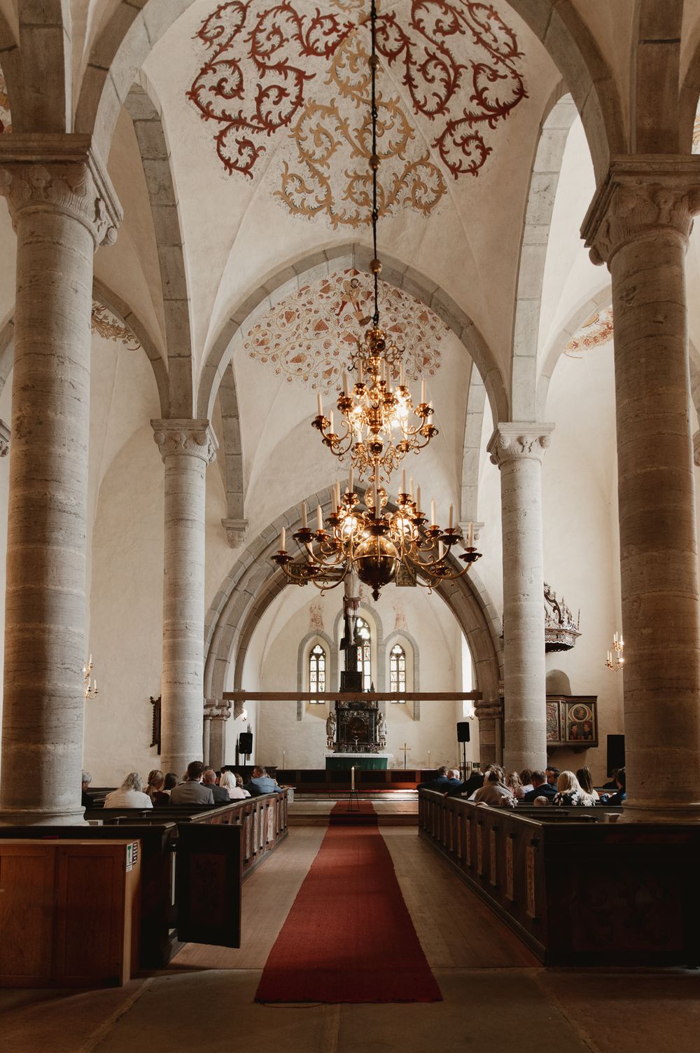 Historic church interior with vaulted ceilings, ornate chandelier, red carpet aisle, and wooden pews filled with visitors.