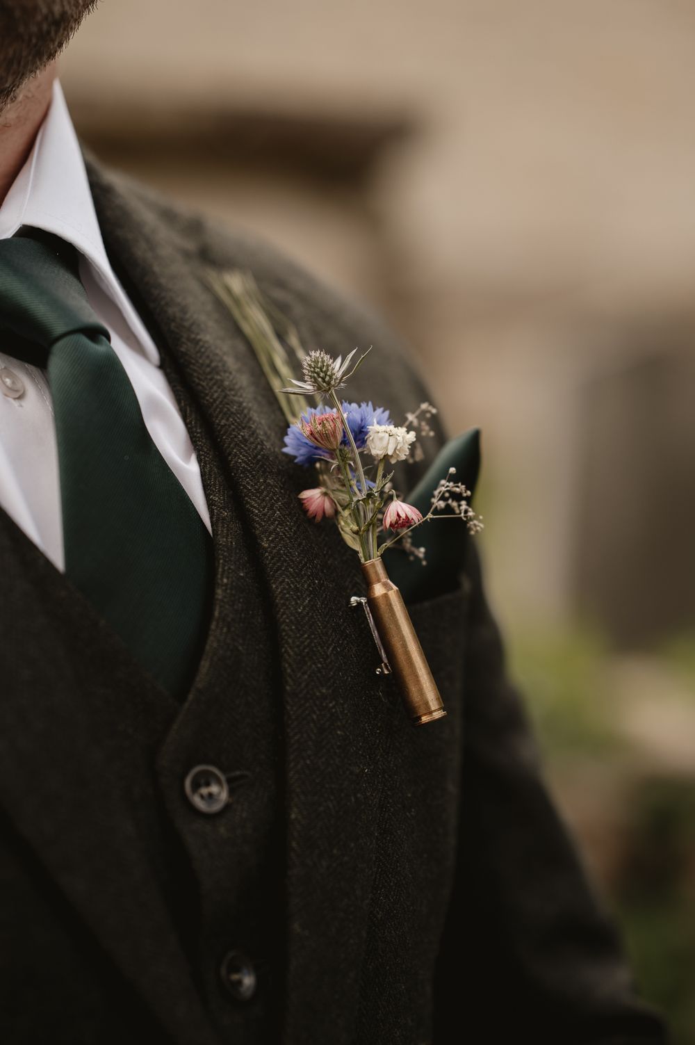 Close-up of a groom wearing a green tweed suit with a wildflower boutonniere in a brass bullet shell vase.