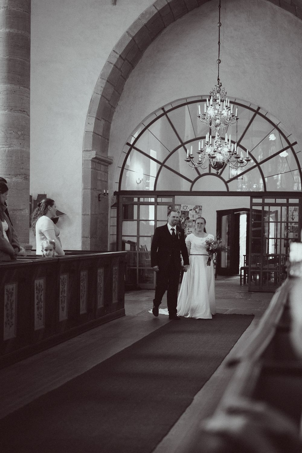 Black and white photo of a bride and groom walking down the aisle of an elegant church with chandelier lighting.
