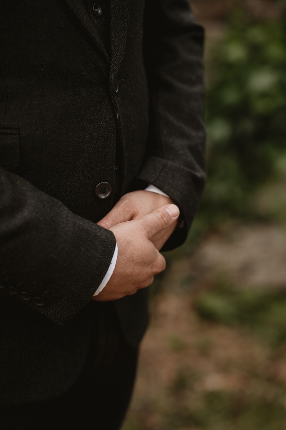 A man in a dark charcoal suit clasps his hands together, wearing a white dress shirt, with a blurred green background.