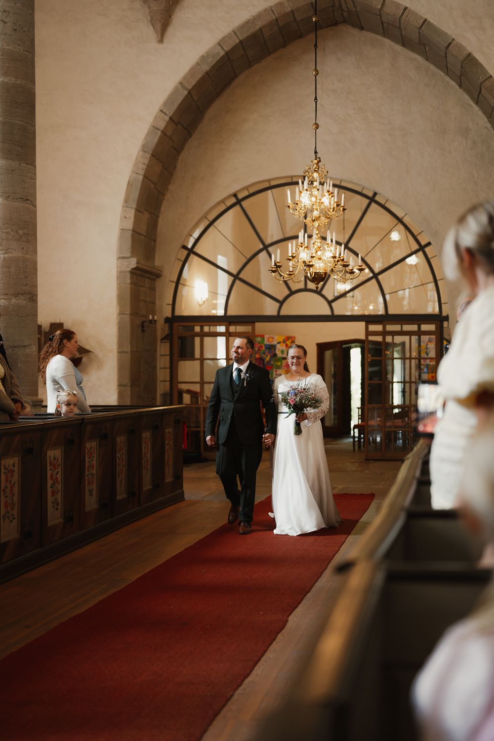 Bride and groom walking down red carpet aisle in historic stone church with arched ceiling and chandelier.