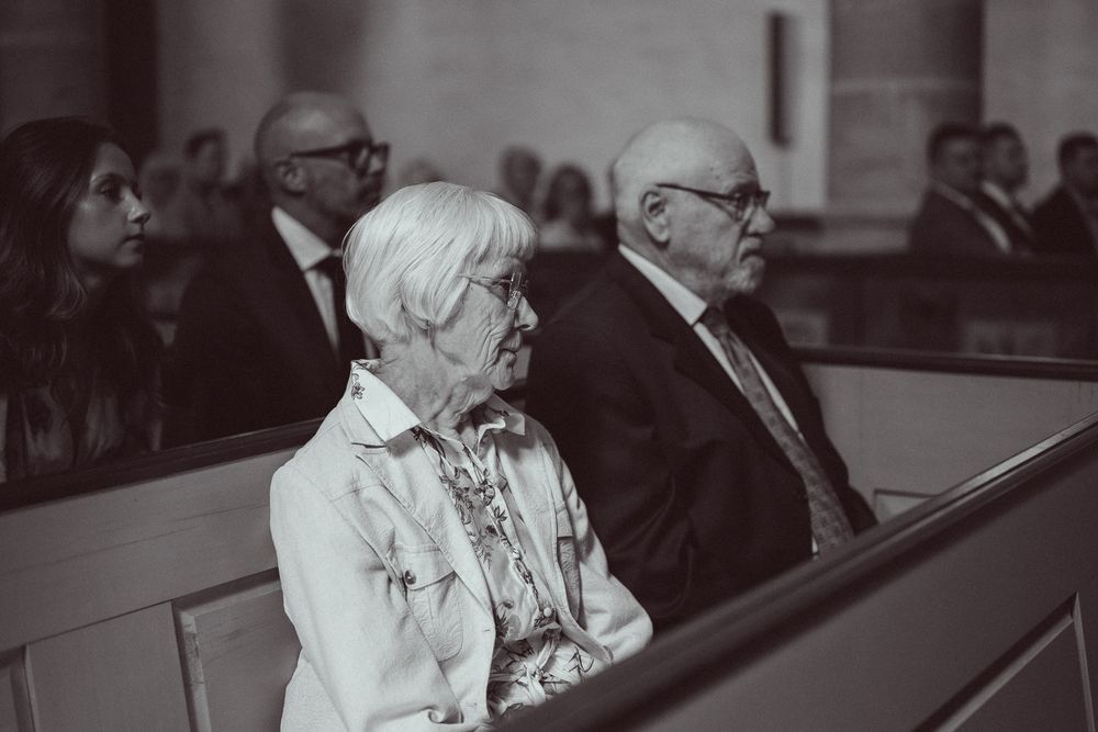 Elderly couple seated in church pews during a formal ceremony, black and white wedding photography.