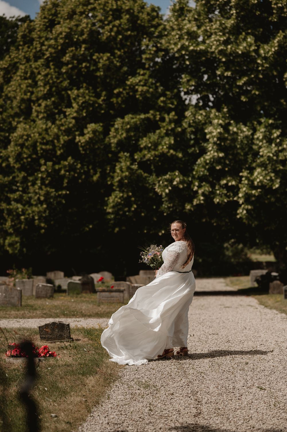 Bride in white lace gown twirling on gravel path in sunlit cemetery garden setting.