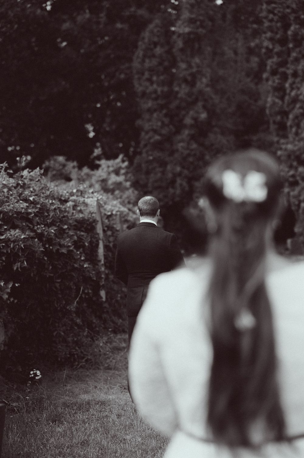 Black and white photo of a bride watching groom walk through lush garden path on wedding day.