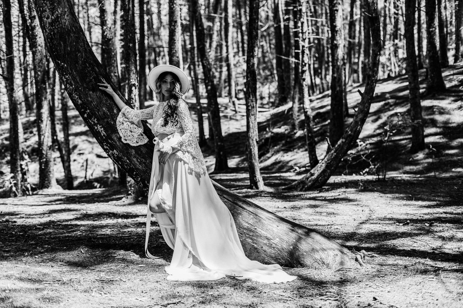 Woman in yellow floral top and black flared pants posing among tall trees in a sunlit forest.