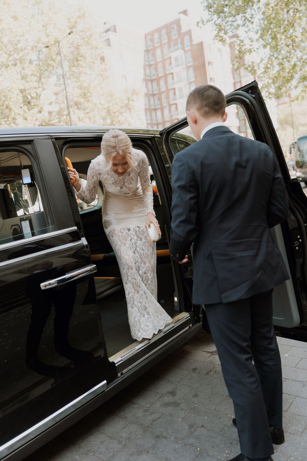 Bride in lace gown stepping out of black limousine, assisted by groom in navy suit on city street.
