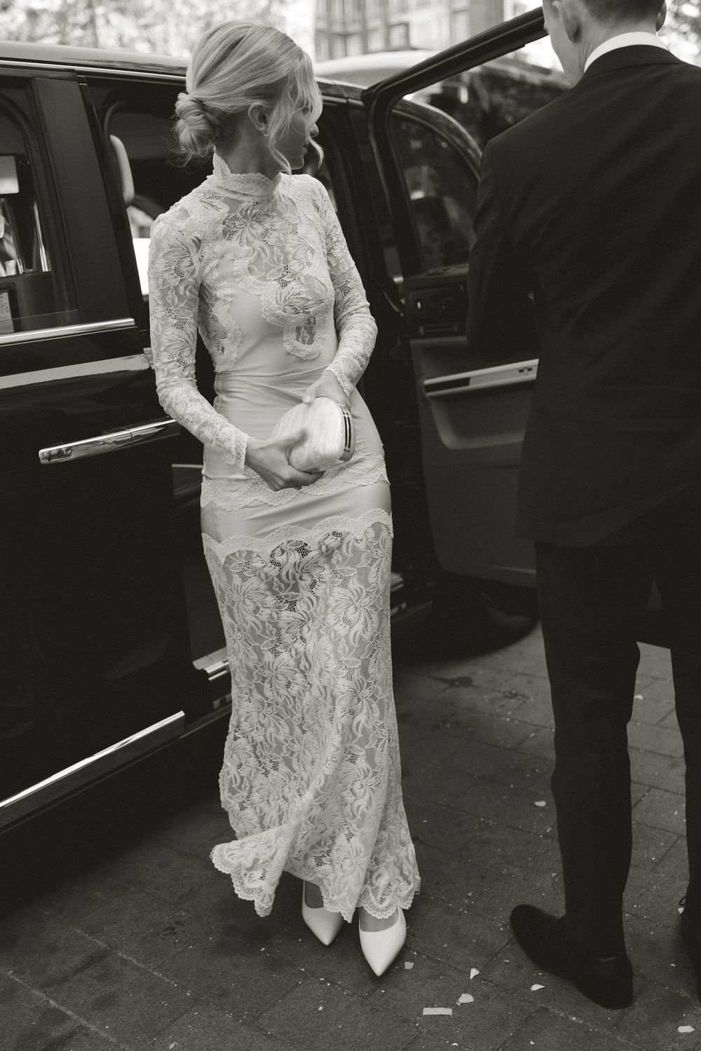 Bride in elegant long-sleeve lace gown holding clutch, stepping out of luxury black car assisted by man in suit.
