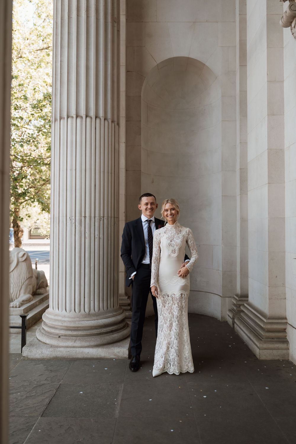 Couple posing in elegant wedding attire between grand stone columns at a classic architectural venue.