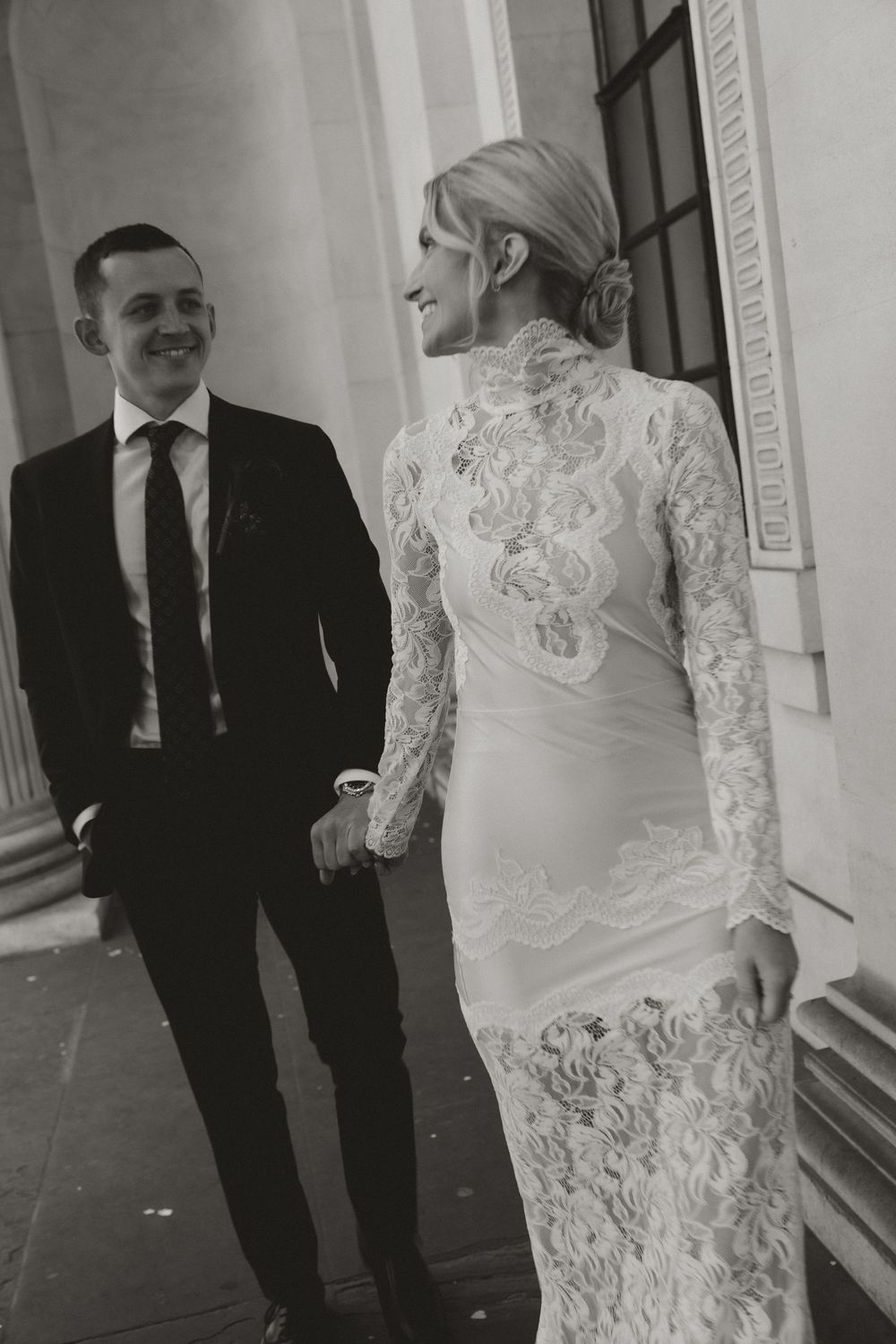 Black and white photo of a bride in lace gown and groom in suit sharing a romantic moment outdoors.