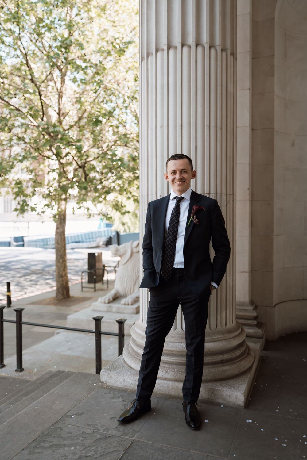 Groom in black suit and tie smiles beside a grand column at a classic architectural wedding venue.