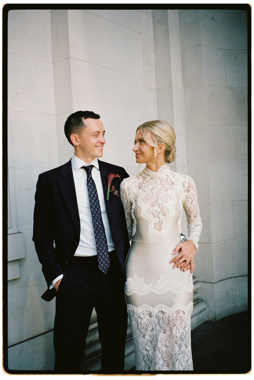 Couple in wedding attire smiling at each other, groom in navy suit, bride in long-sleeve lace dress, standing by stone columns.