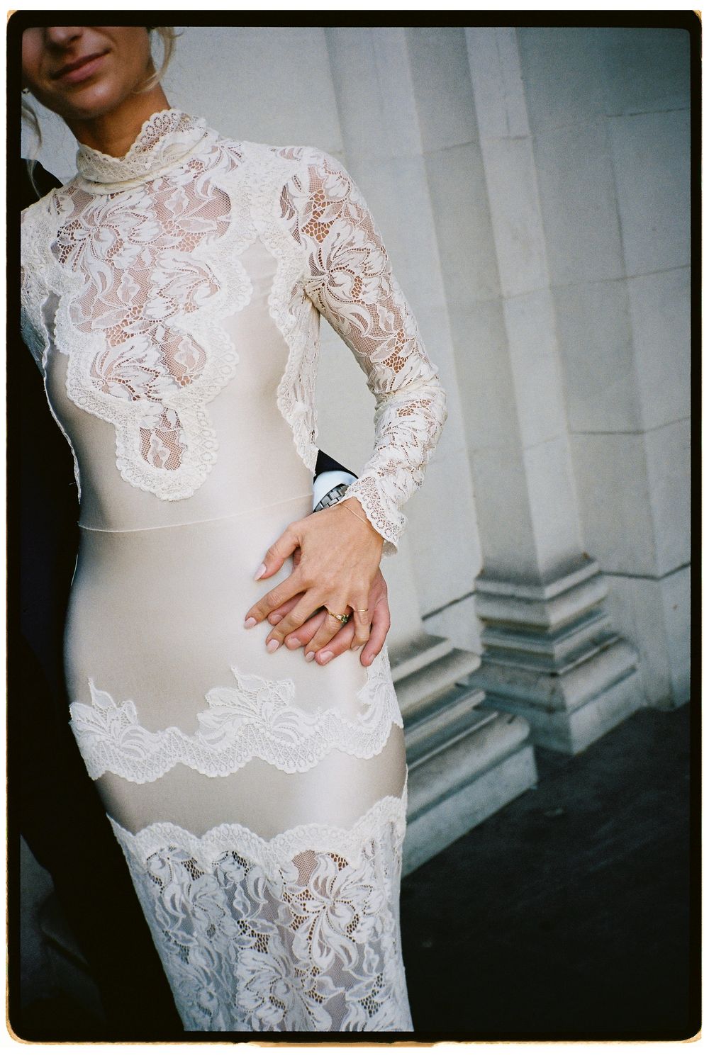 Bride wearing elegant white lace long-sleeve fitted wedding dress with sheer panels, standing beside groom outdoors.