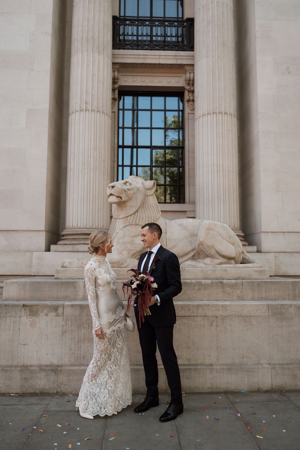 Bride and groom stand before a neoclassical building with a stone lion sculpture, bride in lace gown holding a bouquet.