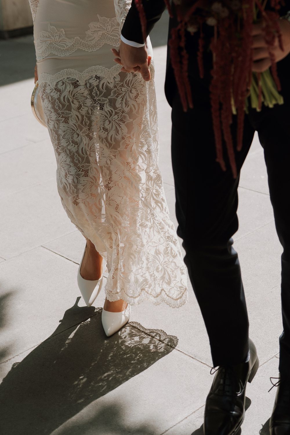 Bride in lace wedding gown and white heels walks beside groom in black suit on sunlit pavement.