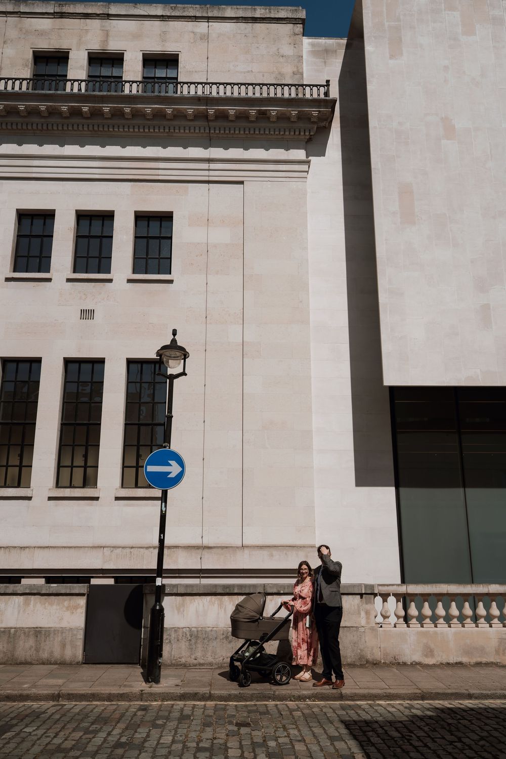 Couple with baby stroller standing on cobblestone street beside classic white stone buildings and a blue arrow sign.