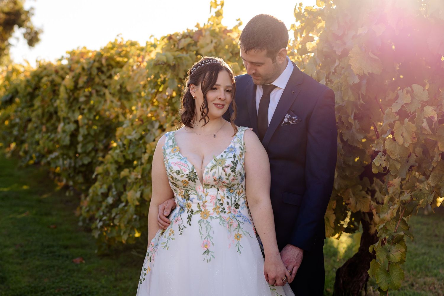 Bride in floral gown and groom in navy suit hold hands at golden hour in a lush vineyard setting.