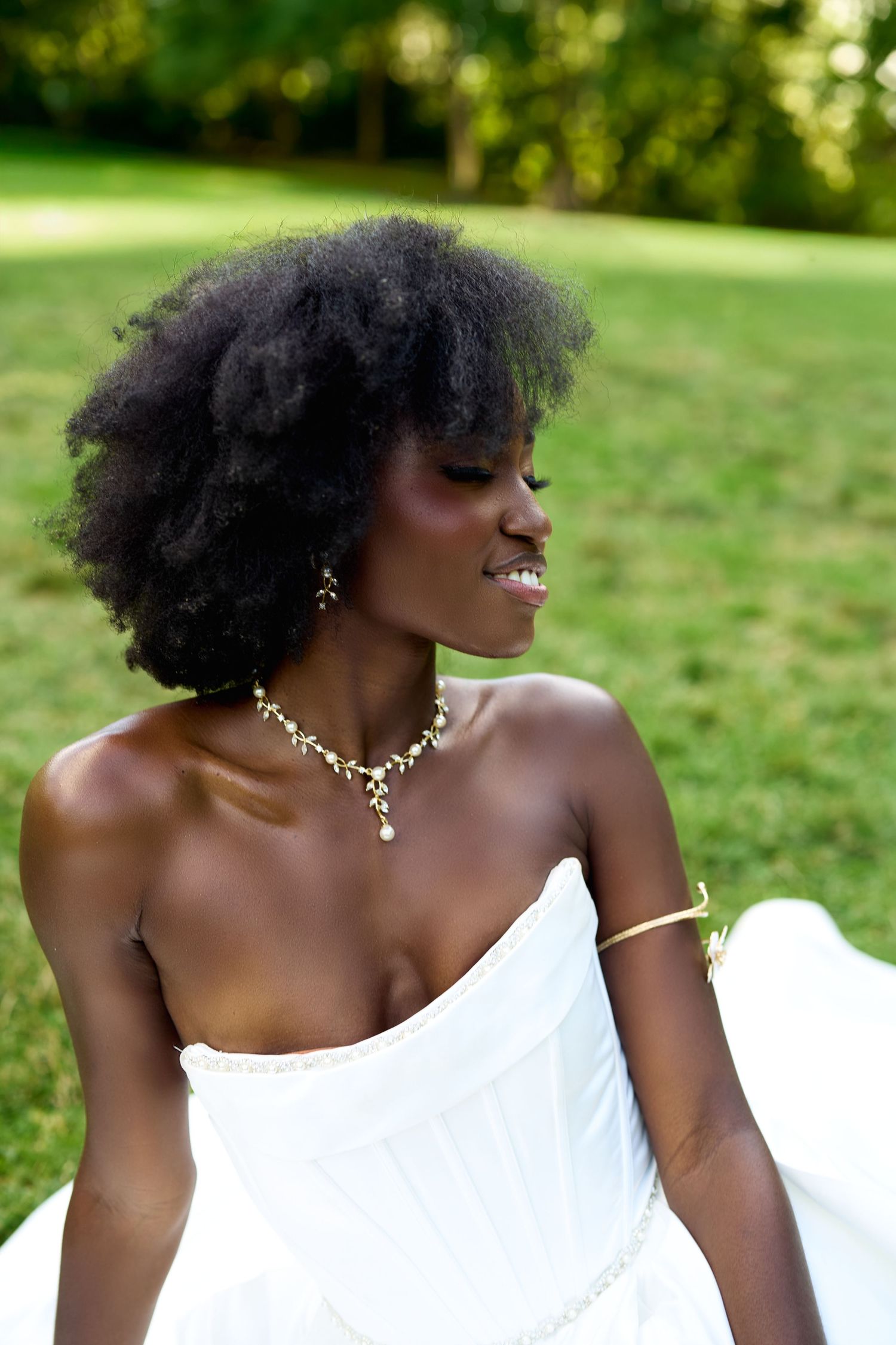 A Black woman in a white strapless dress with gold jewelry and natural afro hair smiles outdoors in a green park.